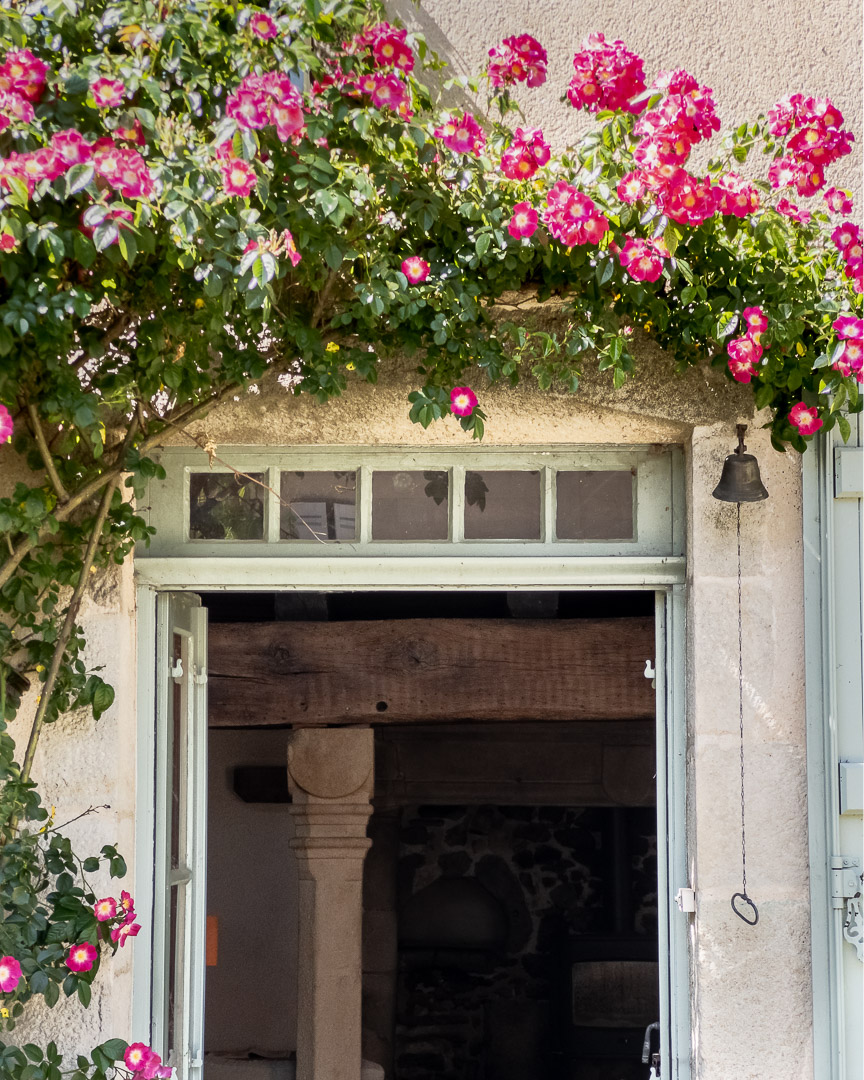 Entrée fleurie d'un ancien presbytère du XIVe siècle avec rosiers, cloche ancienne et vue sur la cheminée à Gourdon près de Cluny.