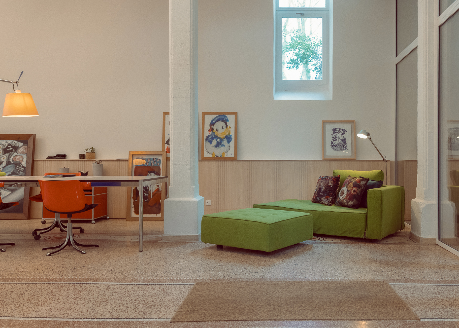 Espace ouvert avec table de travail, fauteuils orange, canapé vert, œuvres d’art et terrazzo dans une ancienne chapelle à Montpellier.